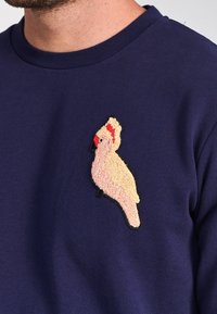Felpa blu navy con un patch ricamato di un cacatua rosa strutturato sul lato sinistro del petto, dotato di una cresta rossa e becco nero.