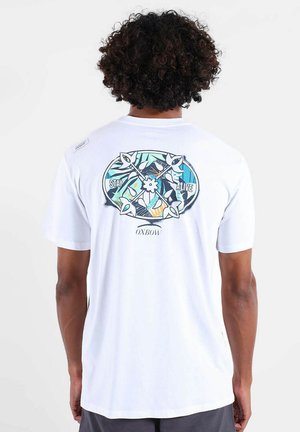 Homme aux cheveux bouclés portant un t-shirt blanc avec des flèches colorées croisées et le texte "Restez en vie" au dos, debout devant un fond blanc.