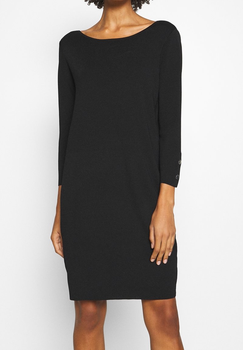 Robe noire en tricot avec un décolleté bateau et des manches trois-quarts. Caractéristiques des accents de bouton sur les poignets. Texture lisse, longueur genou.