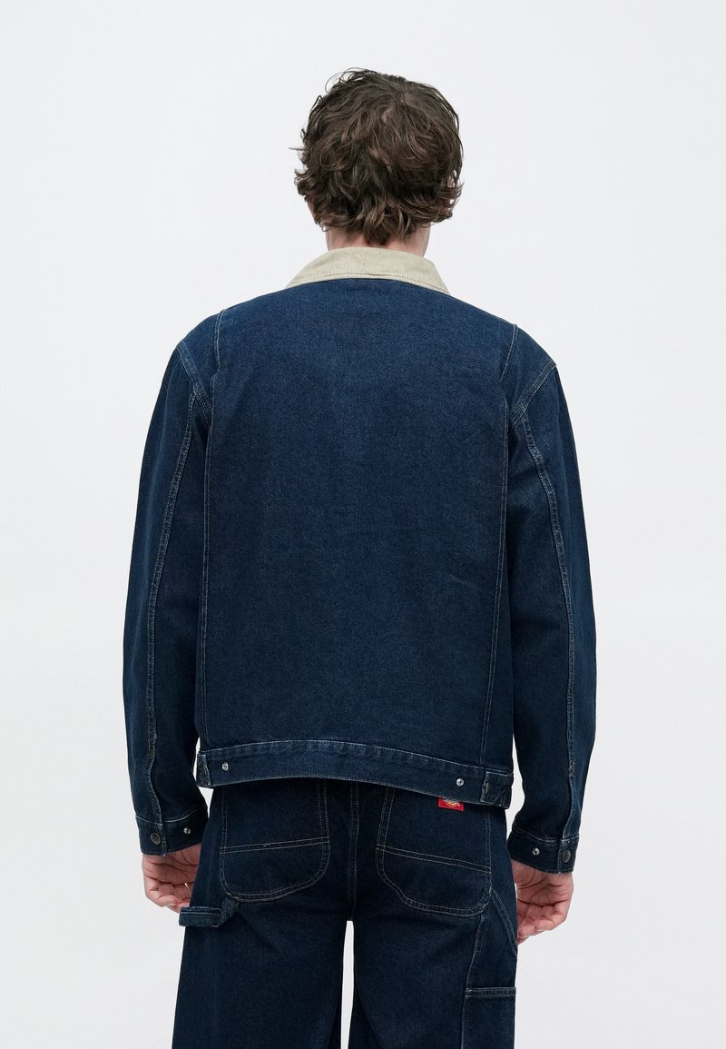Veste en denim bleu foncé avec un col beige contrastant et des coutures apparentes. Présente une coupe droite et une fermeture à boutons à l'ourlet.