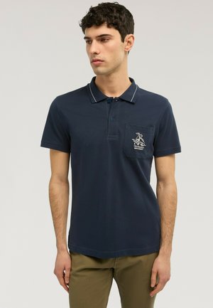 Hombre vistiendo una camisa polo azul marino con un logo blanco en el bolsillo del pecho y pantalones verde oliva, mirando hacia la izquierda contra un fondo liso.