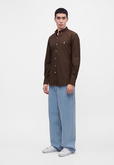 Jeune homme portant une chemise marron foncé boutonnée, un jean bleu clair à jambes larges et des baskets blanches, debout devant un fond uni.