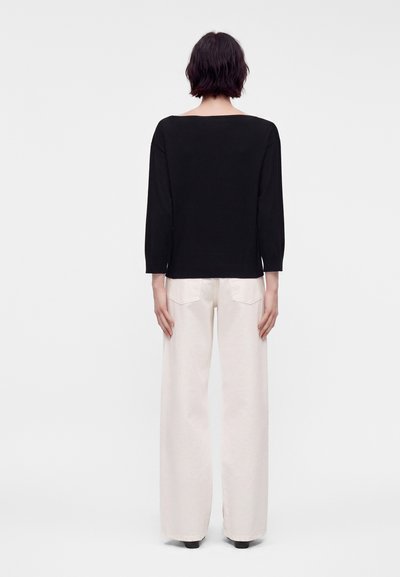 Femme aux cheveux courts et foncés portant un haut noir à manches longues et un pantalon blanc à jambes larges, debout de dos sur un fond uni.