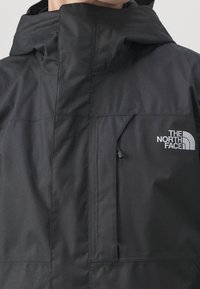 Veste noire à capuche avec un col haut, une poche poitrine zippée et un logo blanc "The North Face" sur la poitrine droite.