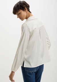 Blusa bianca a maniche lunghe con collo, texture liscia, fessure laterali e piega centrale sul retro, abbinata a jeans di denim blu.