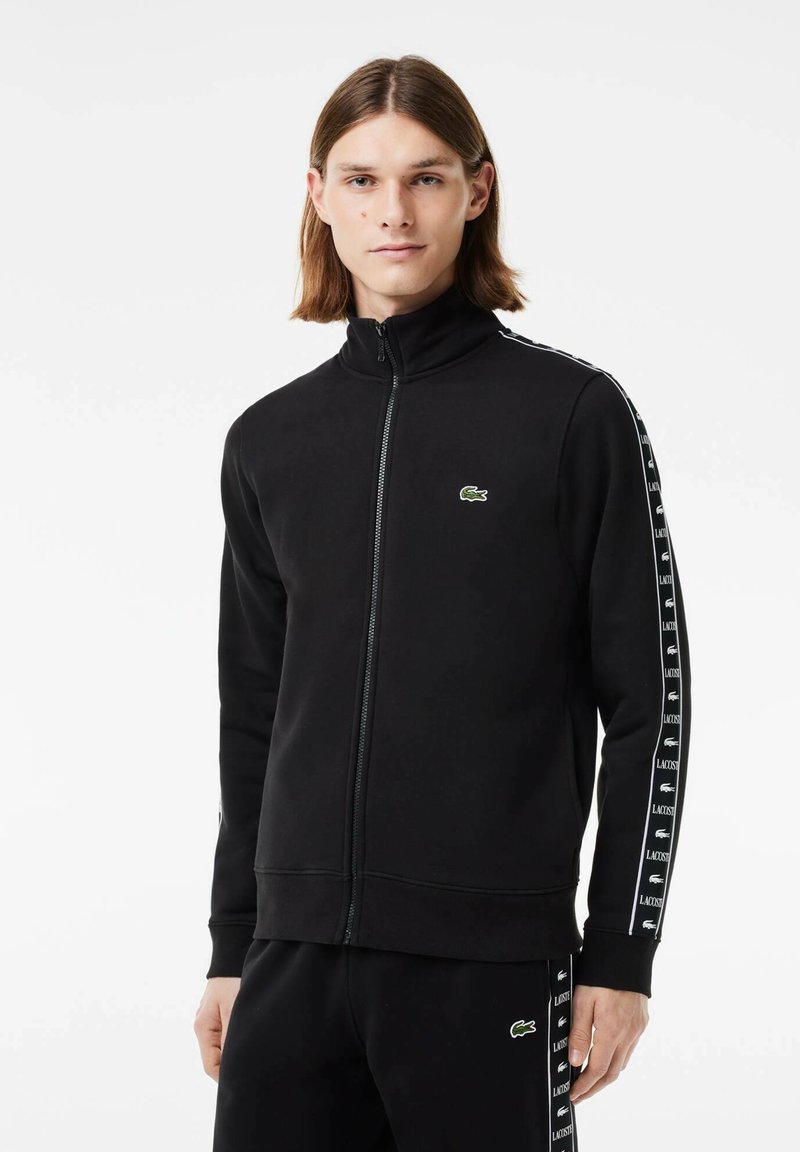Schwarzer Reißverschluss-Sweatshirt mit hohem Kragen, mit weißen Seitenstreifen, die mit "LACOSTE" bedruckt sind, und einem grünen Krokodil-Logo auf der Brust.