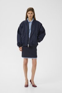 Woman wearing oversized navy bomber jacket over blue polo and pinstriped skirt, paired with burgundy high heels, standing against white background.