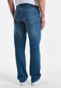 Jeans de mezclilla azul con un corte recto, que cuentan con dos bolsillos traseros, ligero desgaste y una etiqueta en la cintura, combinados con zapatillas blancas.