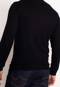 Schwarzer Pullover mit langen Ärmeln, runder Ausschnitt, taillierte Passform und geripptem Saum, kombiniert mit dunkelblauen Jeans aus Denim mit gelben Nähten.