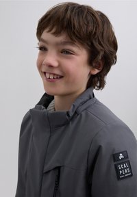 Gray jacket with a high collar, zippered front pocket, and logo patch on the sleeve. Smooth texture, minimal design, tailored fit.