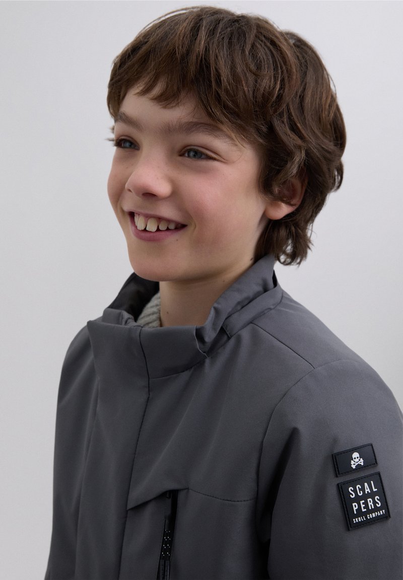 Gray jacket with a high collar, zippered front pocket, and logo patch on the sleeve. Smooth texture, minimal design, tailored fit.