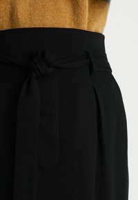 Pantalones negros de talle alto con detalle de cinturón atado, fabricados en una tela suave y con pliegues bien definidos en el frente para añadir textura.