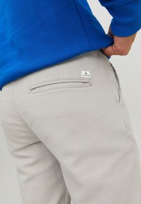 Un pantalon gris clair avec une finition texturée, doté d'une poche arrière avec une étiquette. Stylé avec un sweat-shirt bleu visible en haut.