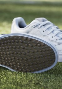 White sneaker with a smooth leather upper, round toe, and flat laces; rubber outsole with textured, wavy pattern. Positioned on green grass.