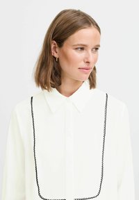 White blouse with a classic collar and black scalloped trim along the front. Smooth texture; lightweight fabric; button closure.