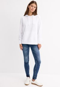Sudadera blanca con un bolsillo frontal y cordón, combinada con jeans skinny de color azul oscuro y zapatillas blancas con una suela marrón.