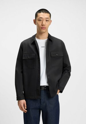 Black button-up jacket with two front pockets, worn over a white t-shirt featuring 'HUGO' logo. Blue denim pants with a black belt.