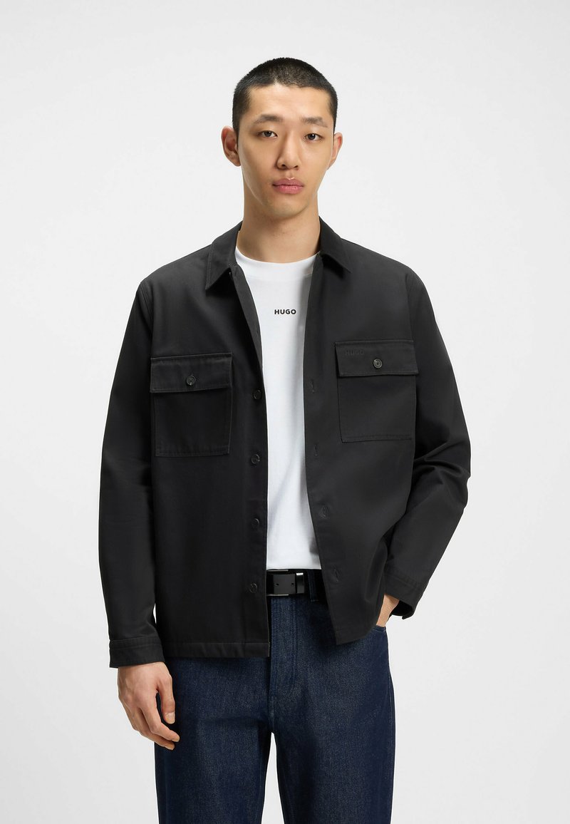 Sort button-up jakke med to frontlommer, båret over en hvid t-shirt med 'HUGO' logo. Blå denimbukser med et sort bælte.
