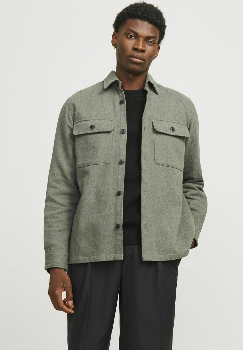 Green wool-blend jacket with two chest pockets, button closure, long sleeves, and a straight cut. Styled over a black sweater, worn with black pants.