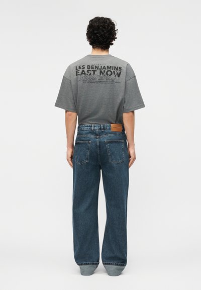 Hombre con cabello rizado que lleva una camiseta gráfica gris y jeans azules holgados, de pie de espaldas contra un fondo blanco liso.