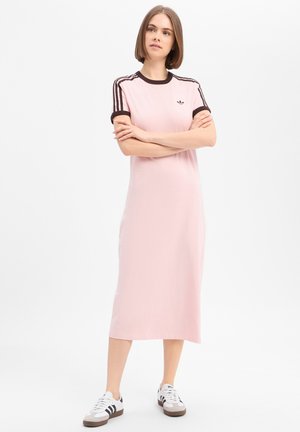 Vrouw staat met armen over elkaar, draagt een roze Adidas midi-jurk met zwarte strepen en witte Adidas sneakers op een witte achtergrond.