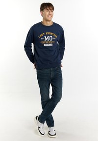Felpa della marina con la scritta "LAST FRONTIER UNIVERSITY" e il grafico "MO" in giallo e bianco. Abbinata a jeans blu e sneaker bianche.