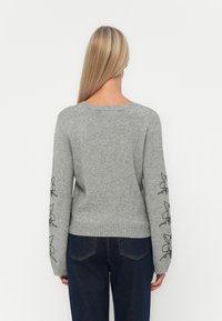 Grå strikkegenser med lange ermer, prydet med svart blomsterbroderi på mansjettene og en ribbestrikket nederkant, designet med rund hals.