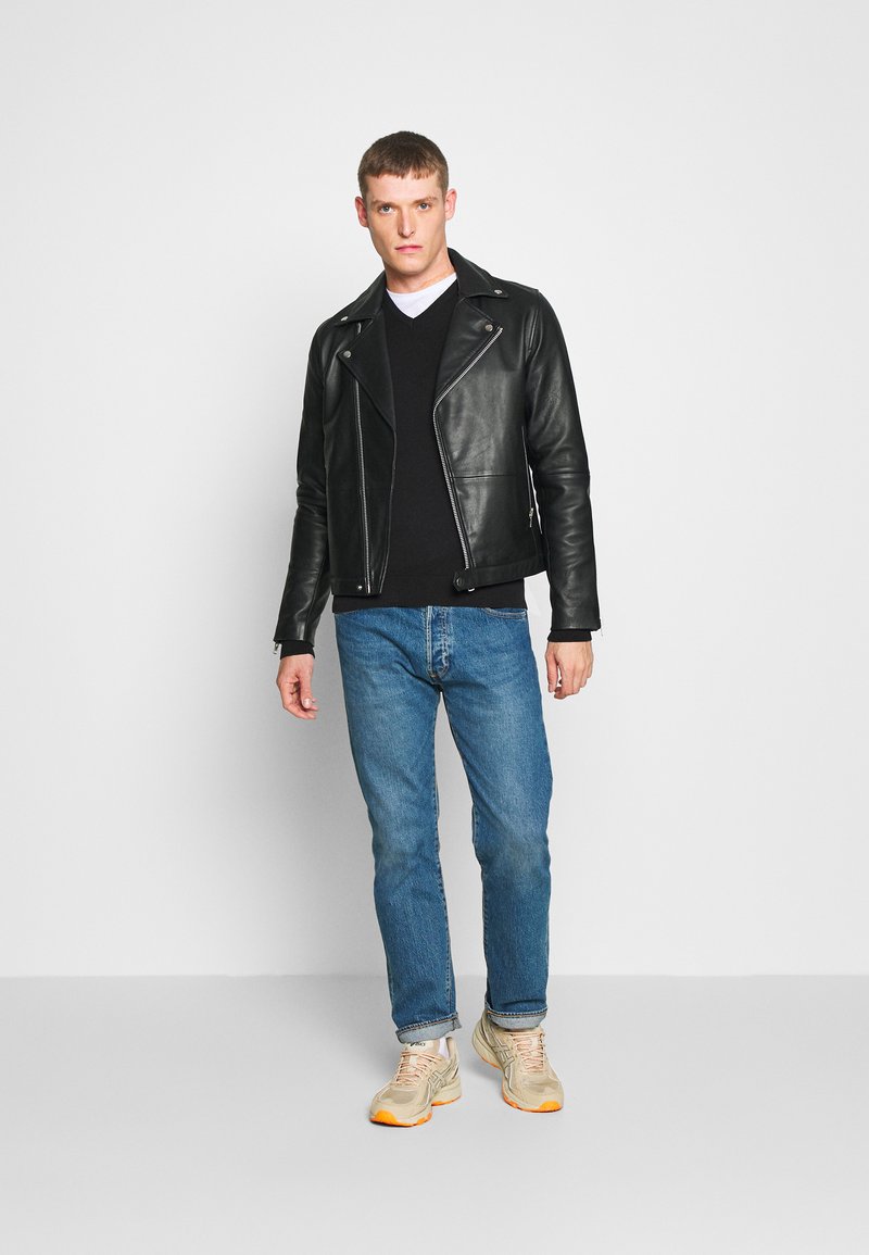 Chaqueta de motorista de cuero negra con cremalleras plateadas, llevada sobre un suéter negro y jeans azules, combinada con zapatillas beige con detalles en naranja.