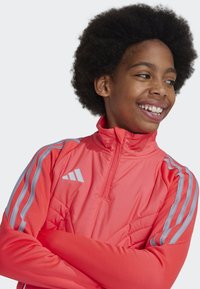 Veste de sport rose vif avec un col haut, fermeture éclair à l'avant et accents rayés gris sur les manches. Présente une texture matelassée et un détail de logo.