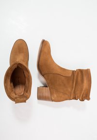 Bruna mockasiner med ankellängd har en rundad tå, samlad skaft och en staplad träklack med vågig sömnadsdetalj längs sidan.