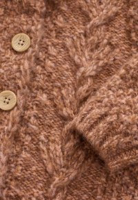 Brązowy sweter dzianinowy z wzorem warkoczowym o miękkiej teksturze. Na przodzie widoczne są dwa drewniane guziki.