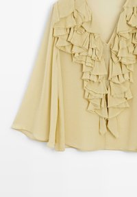 Lysegul bluse med lagdelte flæser langs udskæringen, lange ærmer og et blødt, struktureret stof. V-halsdesign.