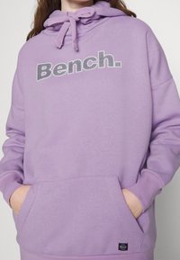 Osoba ubrana w lawendowy hoodie z przednią kieszenią oraz logo "Bench." w białym i czarnym tekście na klatce piersiowej. Ręce w kieszeniach.