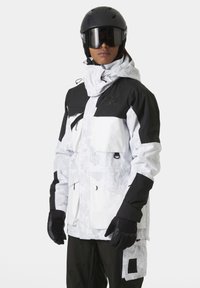 Chaqueta de esquí blanca y negra con múltiples bolsillos, material impermeable y un patrón texturizado. Combinada con guantes negros y un casco.