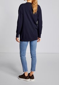 Marinblå tröja med långa ärmar och rundad halsringning, som är kombinerad med ljusblå smala jeans och mönstrade slip-on skor, sedd bakifrån.