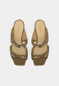 Πέδιλα με ψηλό τακούνι σε χρυσό τόνο, με στριφτά λουράκια, τετράγωνη μύτη και υφή στην επιφάνεια. Το λογότυπο της μάρκας είναι ορατό στην εσωτερική σόλα.