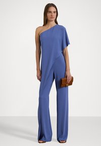 Asymmetrisk en-skulder jumpsuit i blødt blåt stof, der har en afslappet pasform og bred ben-design. Kombineret med en brun clutch.