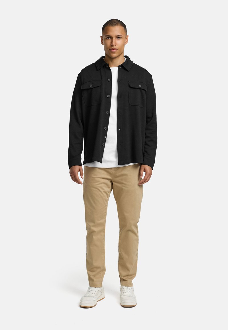 Chemise noire à boutons avec deux poches poitrine, portée sur un t-shirt blanc, associée à un pantalon beige et des baskets blanches. Design minimaliste.