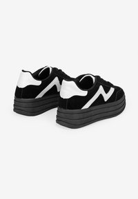 Zapatillas de ante negras con detalles en cuero blanco, que presentan una gruesa suela de goma negra y un diseño en zigzag en el costado.
