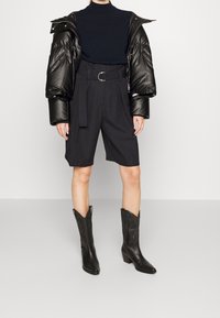 Doudoune noire avec un col haut, associée à un short taillé sombre et des bottes de cow-boy hautes jusqu'au genou avec des détails en relief.