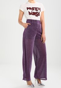 Purple, wide-leg, glittery pants paired with a white t-shirt featuring red graphic text. Model stands with one hand in the pocket. Silver heels.