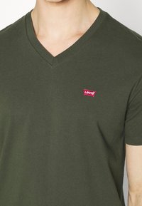 Olivově zelené V-krkové tričko z měkké bavlny, s červeným logem Levi's na levém prsu. Krátké rukávy a volný střih.