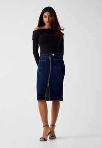 Jupe en denim foncé à silhouette cintrée, longueur genou, avec poches avant, fermeture éclair et surpiqûres contrastées. Le modèle porte un haut noir à épaules dénudées.