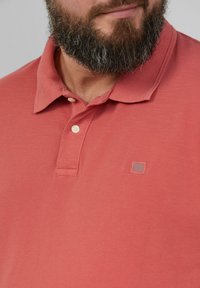 Koronkowa koszulka polo w kolorze koralowym, wykonana z fakturowanej tkaniny, z klasycznym kołnierzykiem, trzema guzikami oraz małym logo na lewej piersi.