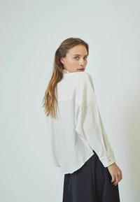 White, textured blouse with a relaxed fit, long sleeves, and buttoned cuffs, paired with a dark skirt. Casual, layered design detail at the back.