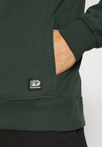 Mano en el bolsillo de un sudadera de Petrol Industries de color verde oscuro, mostrando el puño acanalado y el parche del logo cerca del dobladillo.