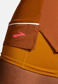 Gros plan d’un short de sport marron et orange avec un logo rose sur la poche latérale et une bande réfléchissante au-dessus.