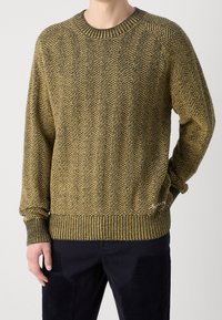 Sweter dziergany w oliwkowym zielonym i żółtym wzorze jodełki, z prążkowanymi mankietami i obszyciem. Posiada małe logo w prawym dolnym rogu.
