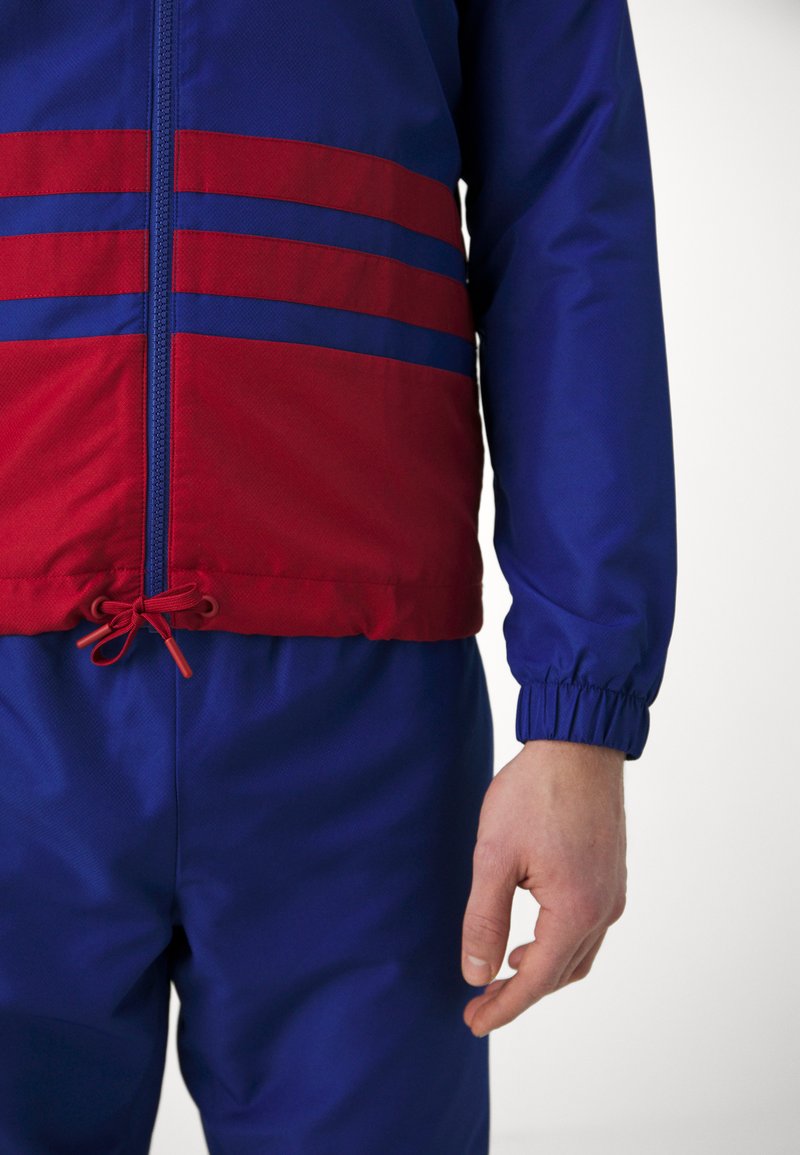 Primo piano di una persona che indossa una giacca a zip blu e rossa abbinata a pantaloni blu, mostrando il polsino elastico della giacca e l'orlo con cordino.