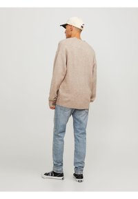 Uomo che indossa un maglione beige, jeans azzurri chiari, sneakers bianco e nere e un cappellino da baseball bianco, in piedi di spalle su uno sfondo semplice.